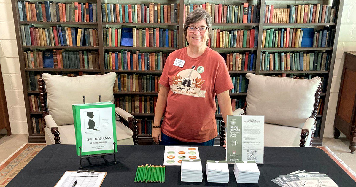 A smiling woman poses with pamphlets, stickers, and pens in front of a large bookshelf.